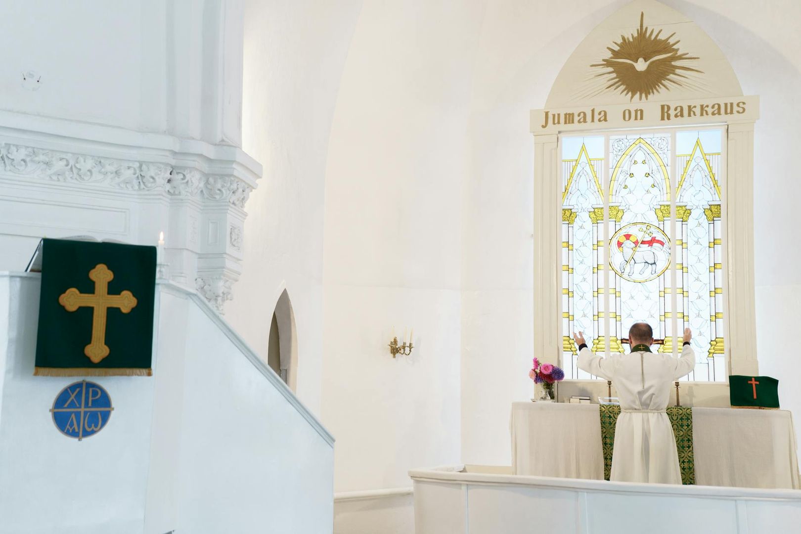 Priester vor Altar mit Buntglasfenster, helle Kirchenwände, kein Akustikpaneel sichtbar.
