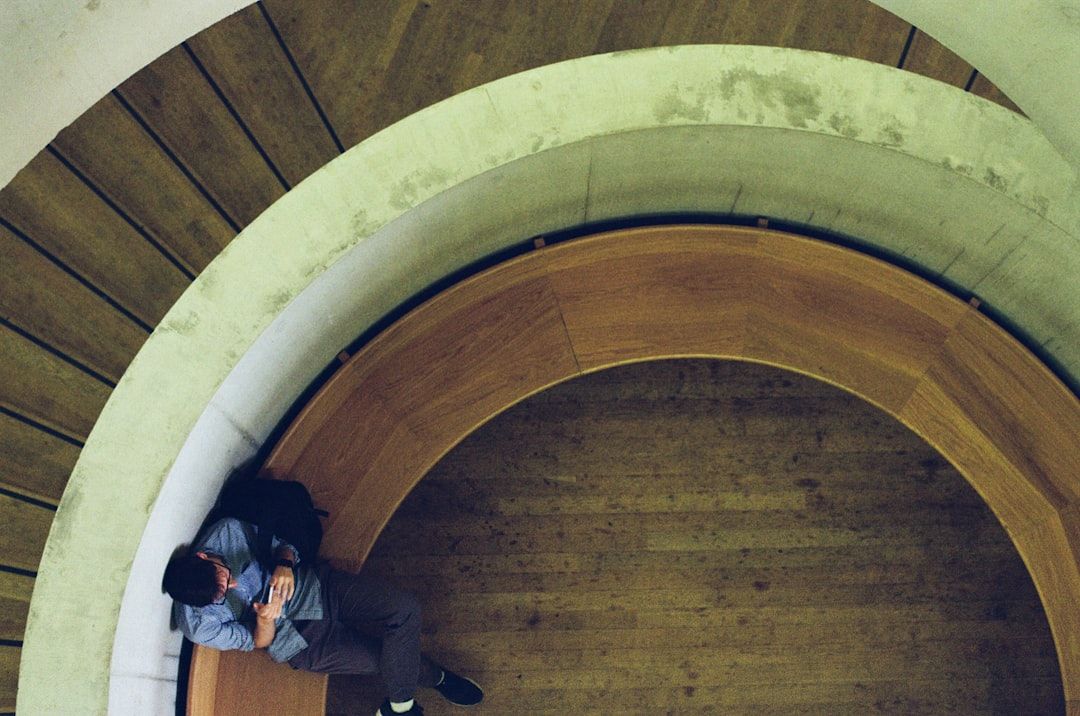Mann sitzt auf einem geschwungenen Holzbänkchen in einem hallenden Treppenhaus.